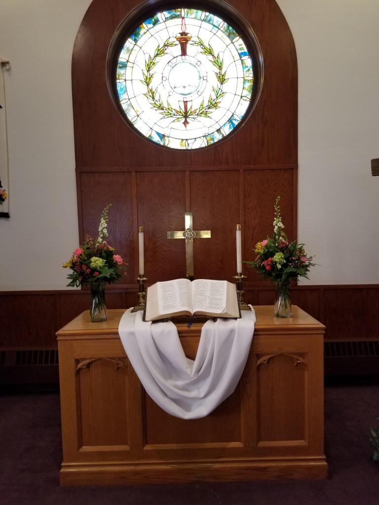 A church alter sits below a circular stained glass window. The window is lit brilliantly from behind with its image of a staff encircled by olive branches very visible. The alter below is decorated with two spring bouqets with bright green, pink, yellow and white flowers. On the alter itself sits two candles on either side of an open Bible and golden cross. A white sash lies beneath all of these things on the table top.
