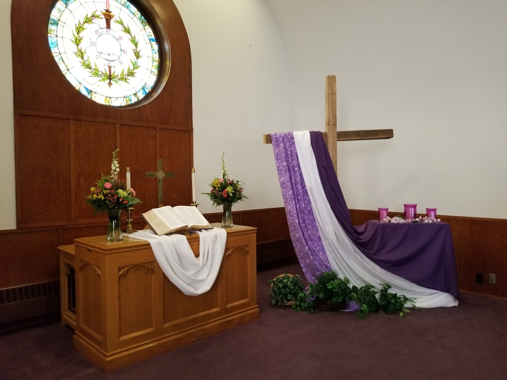An image of the front of a church, with a stained glass window high in the left corner of the window. The stained glass window is lit from behind and the staff and olive branches are clearly visible in it. The alter with flowers, open Bible, candles, white sash, and cross are beneath the window on the lower left corner of the image. On the right half of the image is a large wooden cross, standing taller than the average person. Vibrant purple, violent, and white sashes are draped on the left arm of the cross, running to the floor. A small table sits behind to the right of the cross with another purple sash and some purple items of unknown matter are on top of it.