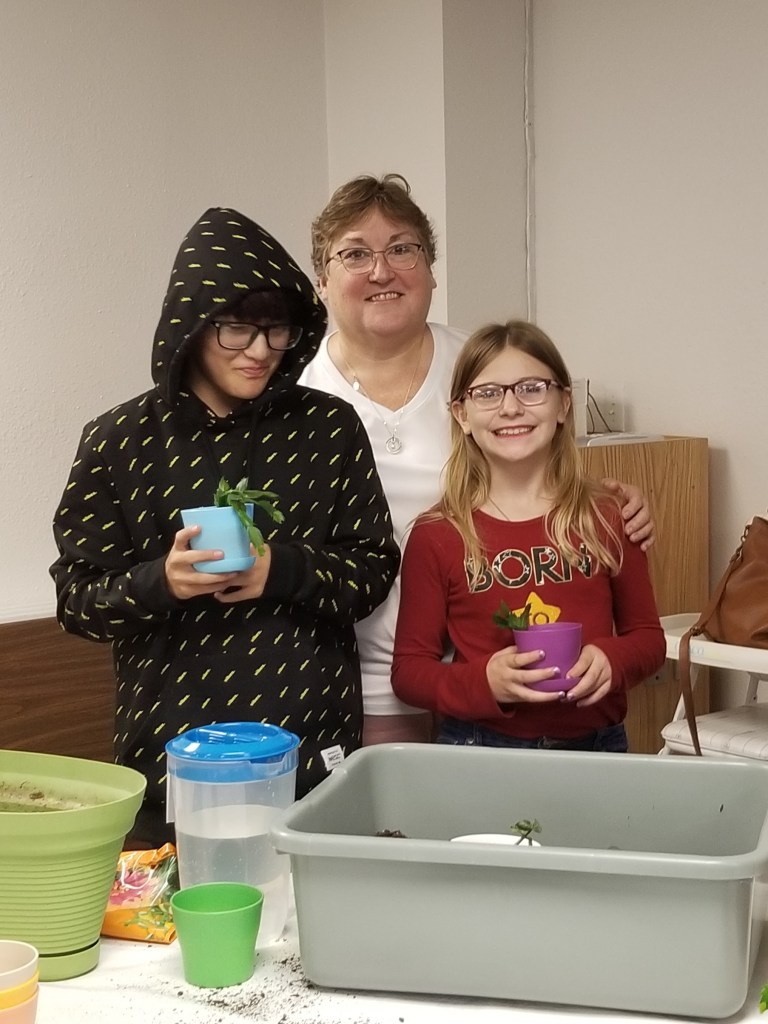 Three people stand against a white wall, with planting materials in front of them. In back is a woman with brown hair and glasses, wearing a white shirt. In front of her are two teens, one on the left wearing a black hoodie zipped up and dark glass and one on the right wearing a red long-sleeved shirt with long blondish hair.