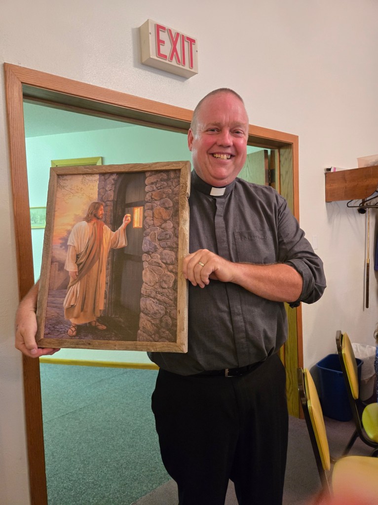 A man in ministerial garb holds up a framed painting of Jesus, knocking at a door.