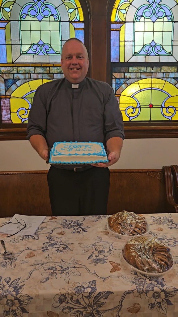 A man in ministerial garb stands in front a stained glass window holding a cake with a message written in frosting. The message is not possible to read from the angle of the photo.